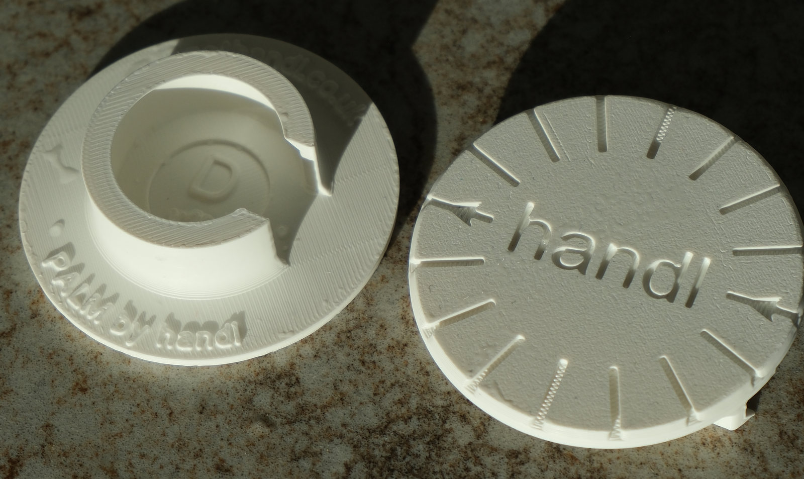 A photograph of two white PALM adapters side by side, one turned each way, so both faces of the part are visible at once. The one on the right is face up: a disc with radial grip grooves around a debossed handl wordmark. The one on the left is face down, showing the spiral channel moulded into the underside — the part that grips the pump collar and holds the disc there — with "PALM by handl" and gethandl.co.uk around its rim.
