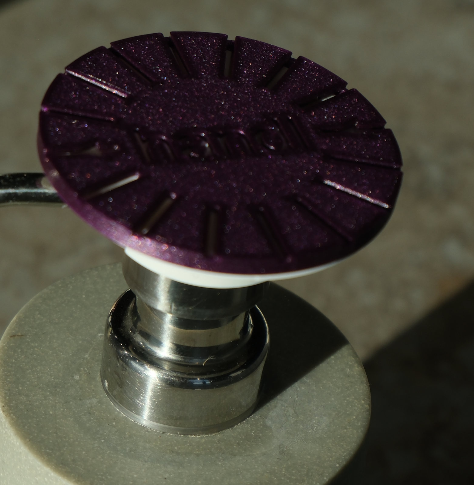 A photograph of a purple PALM clip on a white adapter, the pair seated together on the chrome pump of a plain ceramic soap dispenser. The clip is a broad disc cut with radial grip slots around a debossed handl wordmark; the white adapter beneath it is the part that grips the pump, and between them they turn a narrow pump top into a face wide enough to press with a whole palm.