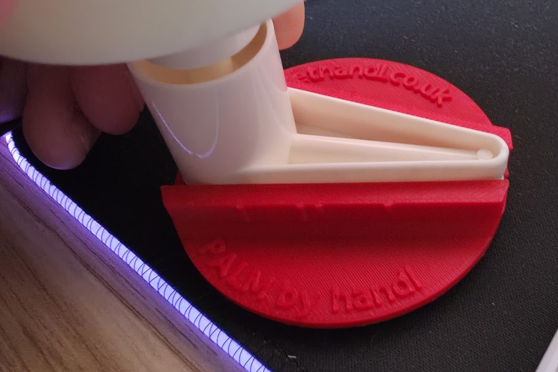 A red 3D-printed PALM disc fitted onto the white L-shaped head of an IKEA TACKAN soap pump, held in one hand. The disc is embossed PALM by handl and gethandl.co.uk.